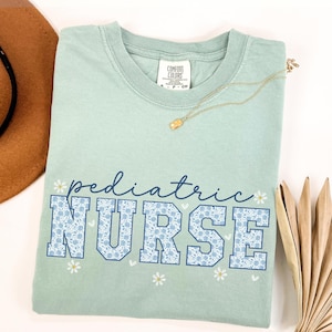 May include: A light blue t-shirt with the text "pediatric NURSE" in blue and white lettering with a floral pattern. The shirt has a white daisy design and a small white heart design.