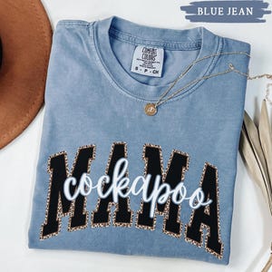 May include: Blue jean colored t-shirt with the words "MAMA cockapoo" in a black and leopard print design. A gold necklace with a pendant is draped across the shirt. The shirt is made by Comfort Colors.
