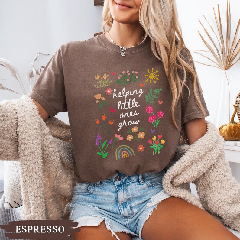 May include: A brown t-shirt with the text "helping little ones grow" surrounded by colorful flowers and a rainbow. The shirt is being worn by a person with long blonde hair and is paired with denim shorts.