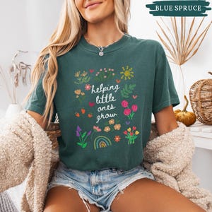 May include: A teal blue t-shirt with a white graphic of flowers, a rainbow, and the text "helping little ones grow".