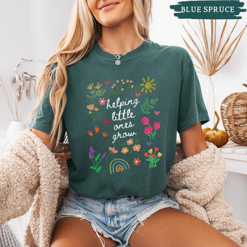 May include: A teal blue t-shirt with a white graphic of flowers, a rainbow, and the text "helping little ones grow".