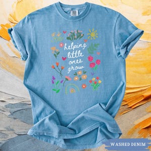 May include: A light blue t-shirt with a white graphic design of flowers and the text "helping little ones grow".