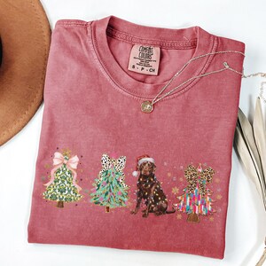 May include: A red t-shirt with a Christmas-themed design featuring three decorated Christmas trees and a chocolate Labrador wearing a Santa hat. The trees are decorated with bows, ornaments, and lights. The text "COMFORT COLORS" is visible on the shirt.