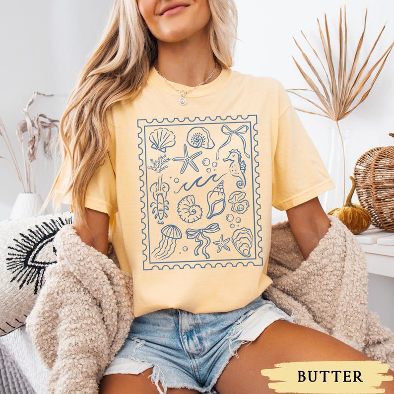 May include: Butter-colored t-shirt featuring a blue stamp design with various sea creatures and elements, including seashells, starfish, and seahorses. The word "BUTTER" is at the bottom of the image.