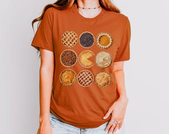 May include: A rust-colored t-shirt with a graphic of nine different pie slices arranged in a grid pattern. The pie slices are various flavors, including pecan, blueberry, and apple. The graphic is reminiscent of the Pac-Man video game.