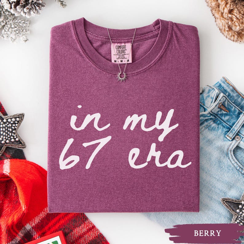 May include: A berry-colored Comfort Colors t-shirt with the phrase "in my 67 era" in white script. A silver necklace with a sunburst pendant is on the shirt. The shirt is folded, with a pair of blue jeans visible.