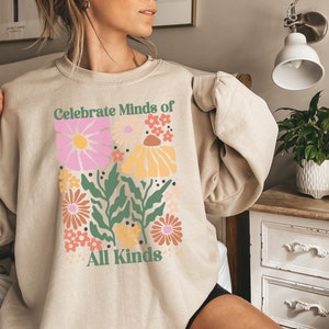 May include: Beige sweatshirt with a floral design and the text "Celebrate Minds of All Kinds." The graphic features pink, yellow, and orange flowers with green leaves. The sweatshirt is a neutral color.