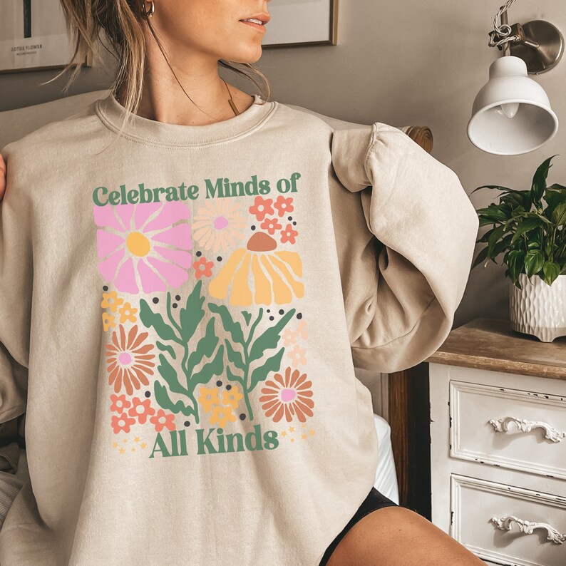 May include: Beige sweatshirt with a floral design and the text "Celebrate Minds of All Kinds." The graphic features pink, yellow, and orange flowers with green leaves. The sweatshirt is a neutral color.