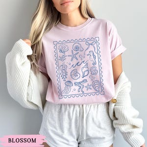 May include: A light pink t-shirt with a blue stamp-like design featuring various sea creatures and elements, including seashells, starfish, and seahorses. The word "Blossom" is printed at the bottom of the image.