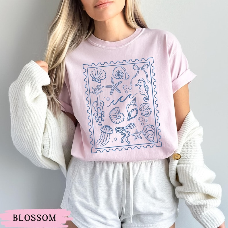 May include: A light pink t-shirt with a blue stamp-like design featuring various sea creatures and elements, including seashells, starfish, and seahorses. The word "Blossom" is printed at the bottom of the image.