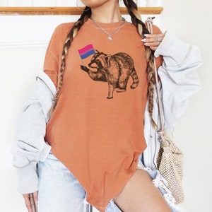 May include: A burnt orange t-shirt with a black and white illustration of a raccoon holding a bisexual pride flag.