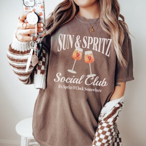 May include: A brown t-shirt with the text "Sun & Spritz Social Club" and the phrase "It's Spritz O'Clock Somewhere" printed in white. The shirt features a graphic of two glasses of Aperol Spritz.