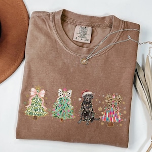 May include: A brown t-shirt with a Christmas tree design featuring a black dog wearing a Santa hat. The trees are decorated with bows, ornaments, and lights. The shirt has the text "COMFORT COLORS" and "S - P - CH" printed on the tag.