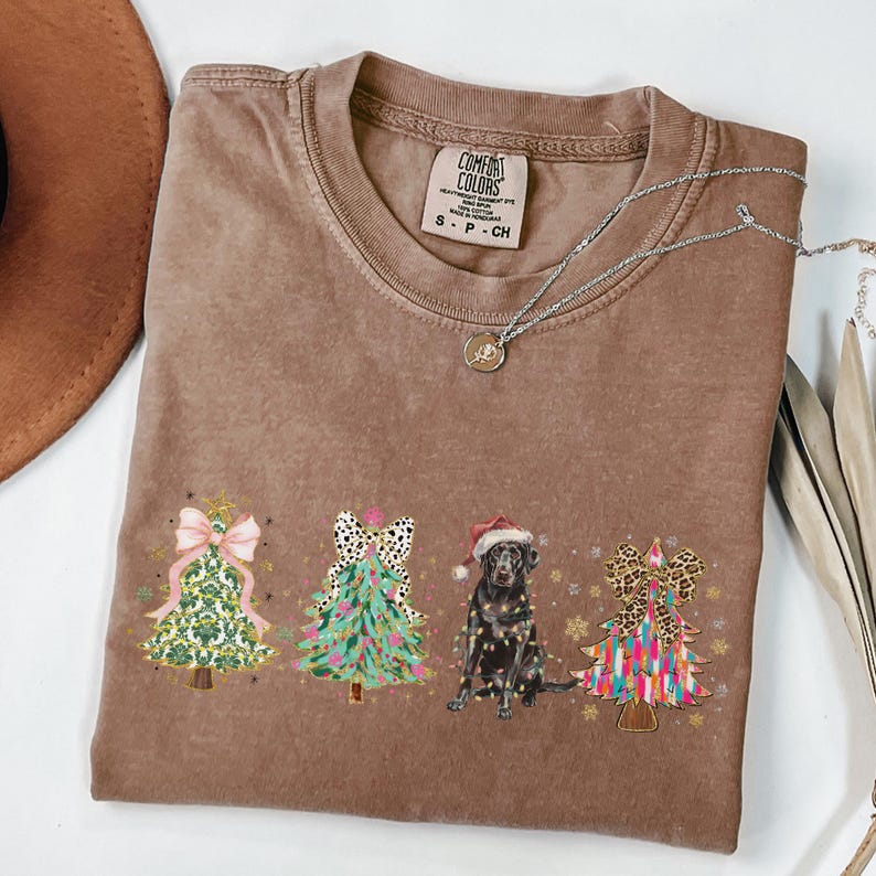 May include: A brown t-shirt with a Christmas tree design featuring a black dog wearing a Santa hat. The trees are decorated with bows, ornaments, and lights. The shirt has the text "COMFORT COLORS" and "S - P - CH" printed on the tag.