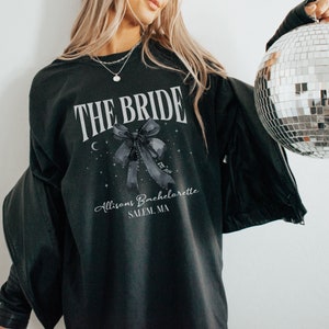K&ouml;nnte beinhalten: Schwarzer Pullover mit dem Text "THE BRIDE" in Wei&szlig;, einer schwarzen Schleife mit wei&szlig;em Band und dem Text "Allisons Bachelorette SALEM, MA" in Wei&szlig;.