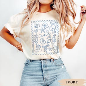 May include: Ivory-colored t-shirt featuring a blue stamp design with various sea creatures and elements, including seashells, starfish, and a seahorse. The shirt's color is labeled "IVORY" at the bottom.
