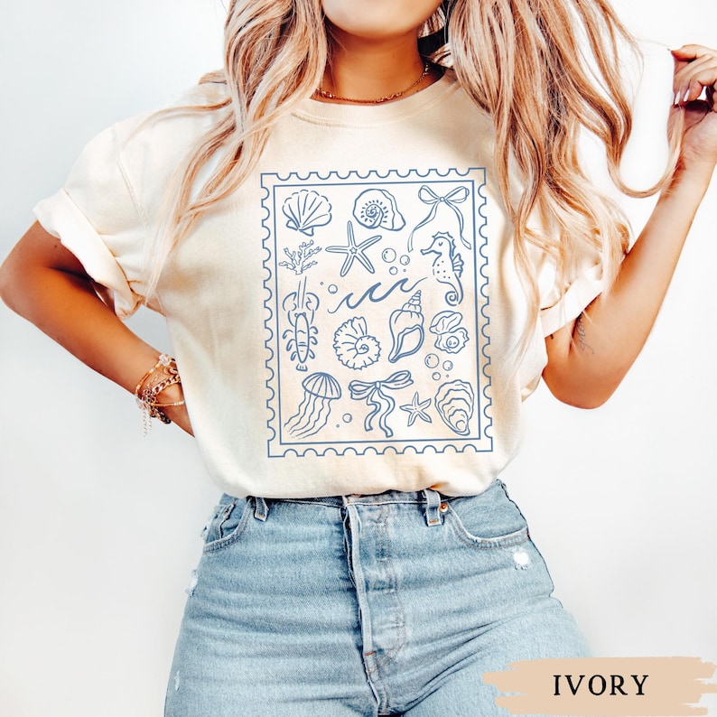May include: Ivory-colored t-shirt featuring a blue stamp design with various sea creatures and elements, including seashells, starfish, and a seahorse. The shirt's color is labeled "IVORY" at the bottom.