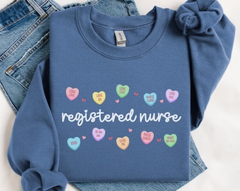 以下が含まれることがあります： 白い文字で「registered nurse」とプリントされた青いスウェットシャツ。テキストは、「code love」、「love dx」、「love stat」、「nurse cupid」、「love rx」などのフレーズが書かれた小さなカラフルなキャンディーハートで囲まれています。