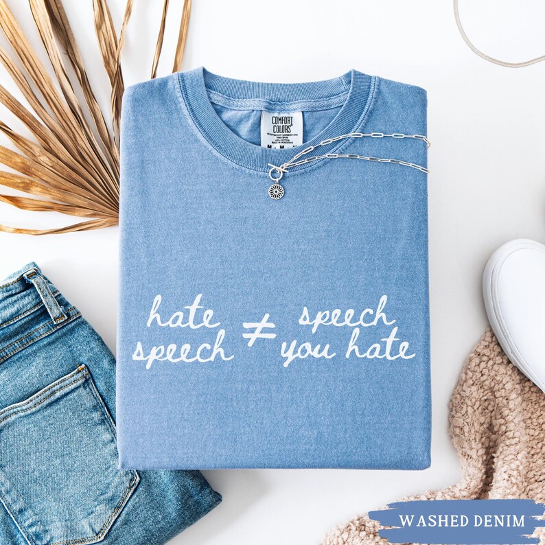 May include: A light blue Comfort Colors t-shirt with the text "hate speech &ne; you hate" in white. A silver necklace with a pendant is on the shirt. The shirt is folded with a pair of blue jeans and a white shoe.