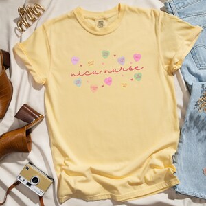 May include: Yellow short-sleeved t-shirt with the text "nicu nurse" in pink script. The shirt is decorated with small pink, yellow, and blue hearts with various sayings on them.