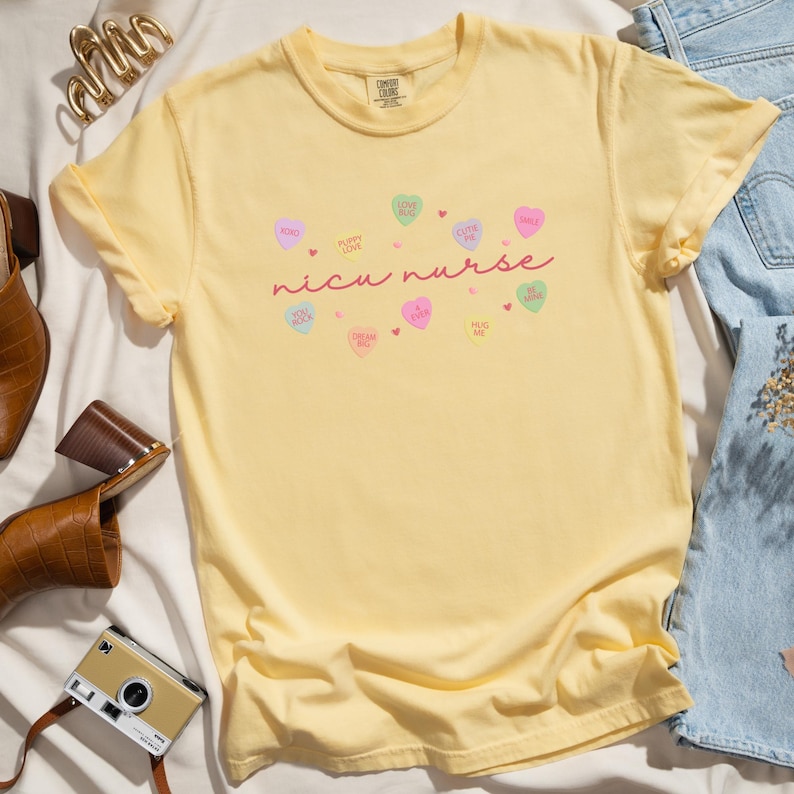 May include: Yellow short-sleeved t-shirt with the text "nicu nurse" in pink script. The shirt is decorated with small pink, yellow, and blue hearts with various sayings on them.