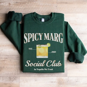 May include: Green sweatshirt with a white graphic of a margarita with the text "Spicy Marg Social Club Est 1983 In Tequila We Trust"