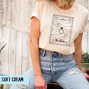 May include: A soft cream t-shirt with a black and white illustration of a hand holding a ring. The illustration is surrounded by a decorative frame and the text "The Bride".