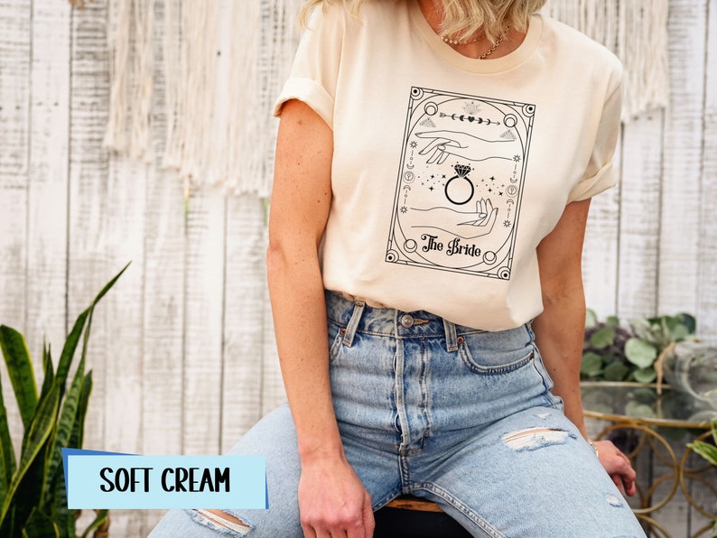 May include: A soft cream t-shirt with a black and white illustration of a hand holding a ring. The illustration is surrounded by a decorative frame and the text "The Bride".