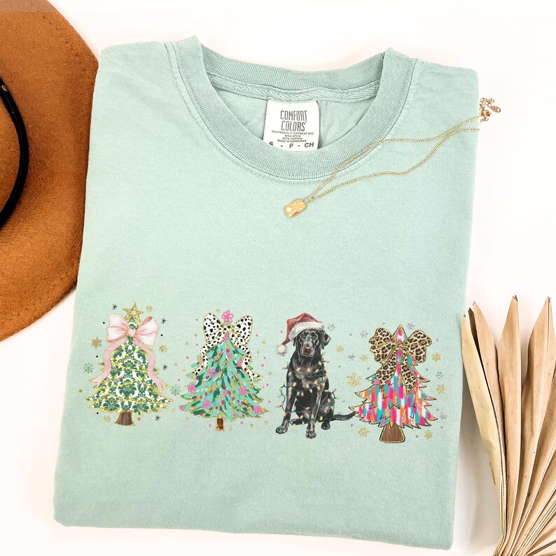 May include: A light green t-shirt with a Christmas tree design featuring a black dog wearing a Santa hat. The trees are decorated with bows, ornaments, and lights. The text "Comfort Colors" is visible on the tag.