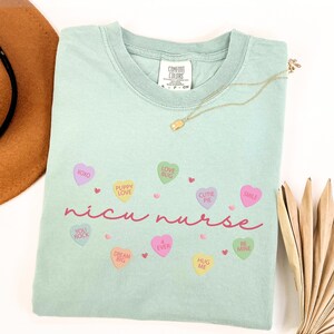 May include: A light green t-shirt with the text "nicu nurse" in pink script. The shirt is decorated with small pink hearts and candy hearts with phrases like "XOXO", "Puppy Love", "Love Bug", "Cutie Pie", "Smile", "You Rock", "Dream Big", "4 Ever", "Hug Me", and "Be Mine".