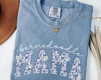 May include: A light blue t-shirt with the text "bernedoodle MAMA" in white. The letters are filled with a floral pattern and paw prints.