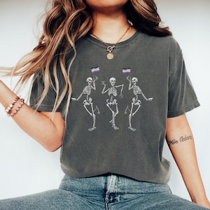 May include: A dark gray t-shirt with a graphic of three skeletons dancing. Each skeleton is holding a small purple, white, and black flag. The text "More Oceans" is visible on the right arm of the model wearing the shirt.