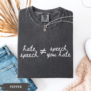 May include: A dark gray Comfort Colors t-shirt with the text "hate speech &ne; speech you hate" in white. A silver necklace with a pendant is draped on the shirt. A pair of blue jeans is visible.