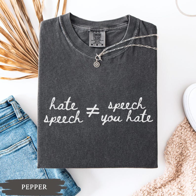 May include: A dark gray Comfort Colors t-shirt with the text "hate speech &ne; speech you hate" in white. A silver necklace with a pendant is draped on the shirt. A pair of blue jeans is visible.