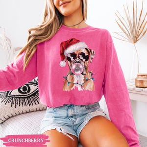May include: A bright pink sweatshirt featuring a Boxer dog wearing sunglasses and a Santa hat, blowing a bubblegum bubble. The dog is adorned with Christmas lights. The word "CRUNCHBERRY" is printed at the bottom.