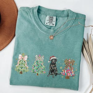 May include: Teal blue t-shirt with a Christmas tree design featuring a black dog wearing a Santa hat and three decorated Christmas trees. The text "COMFORT COLORS" is visible on the tag.