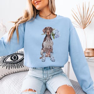 May include: A light blue sweatshirt with a graphic of a brown and white German Shorthaired Pointer dog wearing blue and white rain boots and holding a bouquet of blue hydrangeas in its mouth.