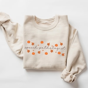 May include: A beige sweatshirt with the text "speech pathologist" in brown script. The text is surrounded by small orange pumpkins and brown leaves.