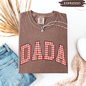 K&ouml;nnte beinhalten: Ein braunes T-Shirt mit dem Wort "DADA" in rot-wei&szlig;em Vichydruck. Das Shirt ist gefaltet und hat eine silberne Halskette mit Anh&auml;nger. Eine Jeans und ein wei&szlig;er Schuh sind sichtbar.