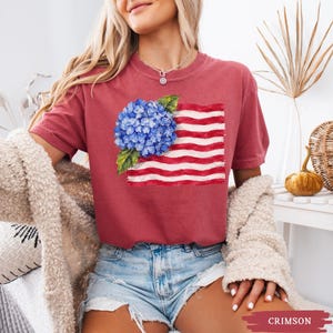 Op de afbeelding: Een karmozijnrood t-shirt met korte mouwen en een grafische print van een blauwe hortensiabloem geplaatst over een gestileerde Amerikaanse vlag met rode en witte horizontale strepen, voorzien van een ronde halslijn.