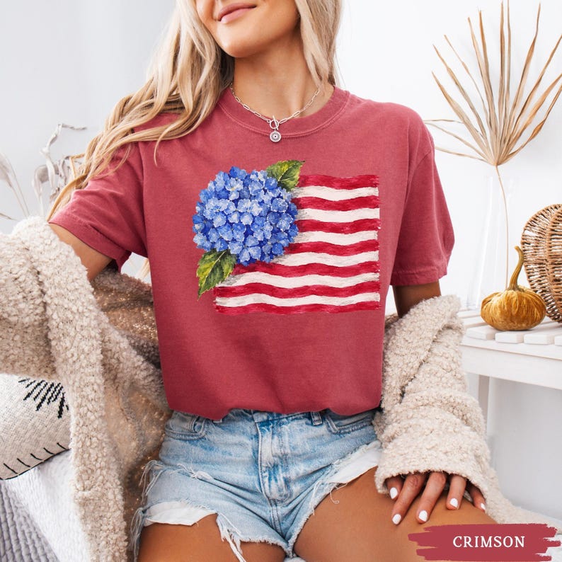 Op de afbeelding: Een karmozijnrood t-shirt met korte mouwen en een grafische print van een blauwe hortensiabloem geplaatst over een gestileerde Amerikaanse vlag met rode en witte horizontale strepen, voorzien van een ronde halslijn.