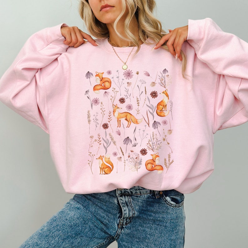 May include: A pink sweatshirt with a graphic print of orange foxes and purple flowers. The sweatshirt has a crew neck and long sleeves.