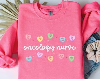 以下が含まれることがあります： ピンクのトレーナーに「oncology nurse」の文字と、さまざまな言葉が書かれたカラフルなキャンディーハートの模様がプリントされています。