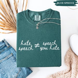 May include: A folded blue spruce Comfort Colors t-shirt with the text "hate speech &ne; you hate" in white script. A silver necklace with a sun pendant rests on the shirt. A pair of blue jeans and a white shoe are in the background.