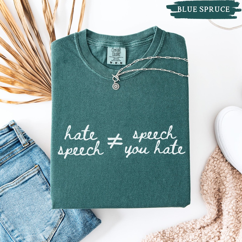 May include: A folded blue spruce Comfort Colors t-shirt with the text "hate speech &ne; you hate" in white script. A silver necklace with a sun pendant rests on the shirt. A pair of blue jeans and a white shoe are in the background.