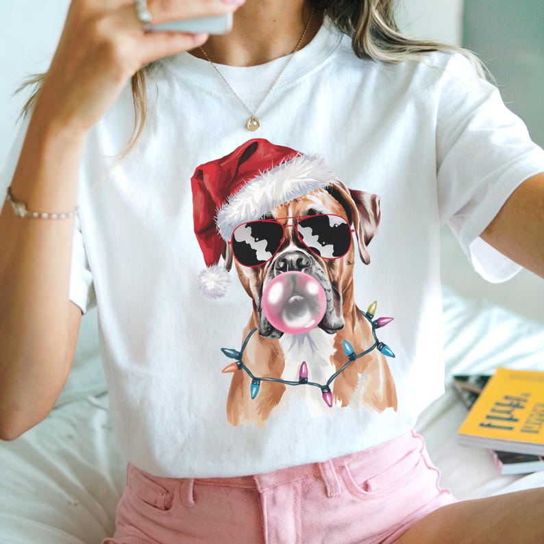 May include: White t-shirt featuring a Boxer dog wearing a Santa hat and sunglasses, blowing a bubblegum bubble. The dog is adorned with a string of colorful Christmas lights. A fun, festive design.