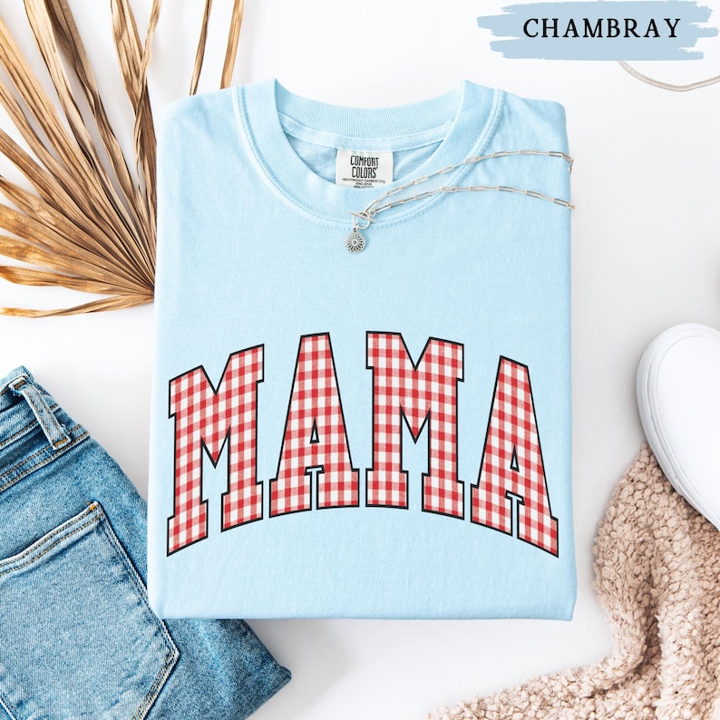 K&ouml;nnte beinhalten: Hellblaues "Comfort Colors" Sweatshirt mit dem Wort "MAMA" in rot-wei&szlig; karierten Blockbuchstaben. Eine silberne Halskette mit einem kleinen Anh&auml;nger befindet sich auf dem Shirt. Das Wort "CHAMBRAY" steht dar&uuml;ber. Eine blaue Jeans und ein wei&szlig;er Schuh sind im Hintergrund.