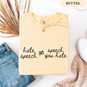 May include: A butter-colored sweatshirt with the text "hate speech &ne; you hate" in black script. The shirt is folded and placed on a white surface next to a pair of blue jeans and a white shoe. A gold necklace is draped on the shirt.