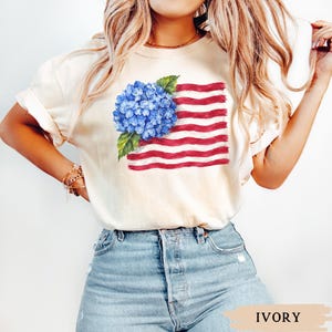 Op de afbeelding: Een ivoorkleurig T-shirt met korte mouwen en een grafische print van een blauwe hortensia-bloemtros geplaatst over een gestileerd, golvend ontwerp van de Amerikaanse vlag met rode en witte strepen, voorzien van een ronde hals.