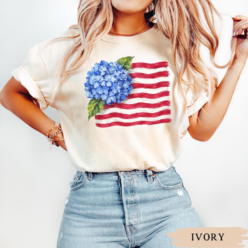 Op de afbeelding: Een ivoorkleurig T-shirt met korte mouwen en een grafische print van een blauwe hortensia-bloemtros geplaatst over een gestileerd, golvend ontwerp van de Amerikaanse vlag met rode en witte strepen, voorzien van een ronde hals.